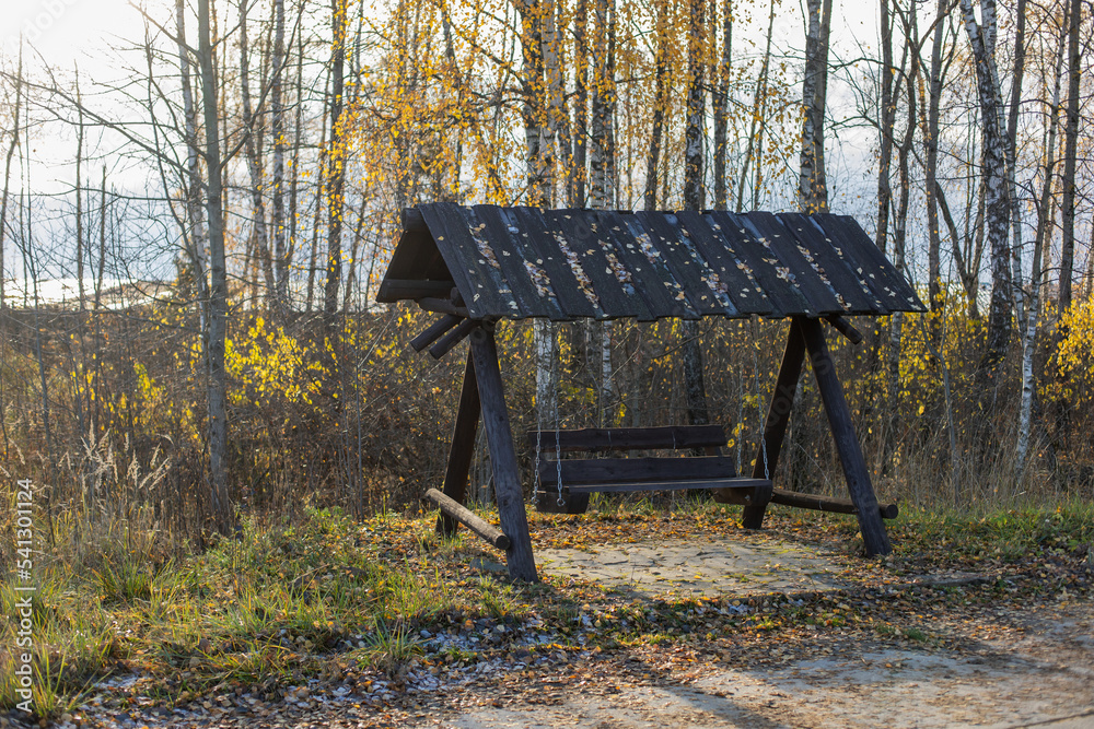 black wooden swing with a large seat in autumn stands by the path with trees in the village. black wooden swing with a large seat in autumn stands by the path with trees in the village.