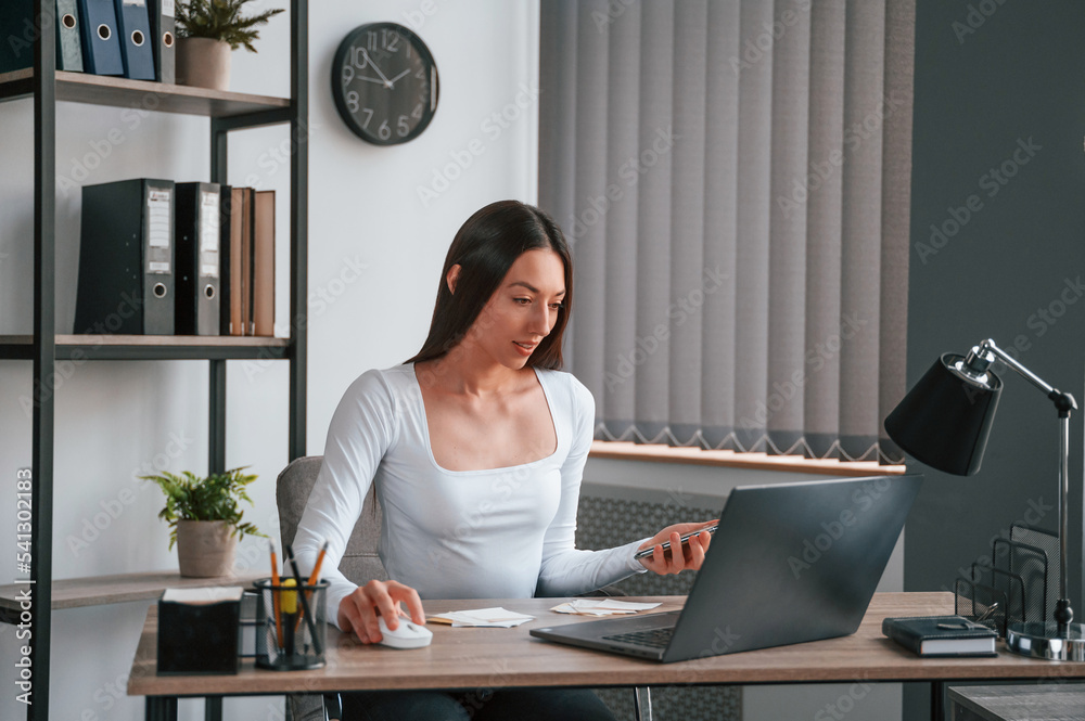 With laptop. Sitting and working. Young beautiful woman in white clothes is in the office With laptop. Sitting and working. Young beautiful woman in white clothes is in the office