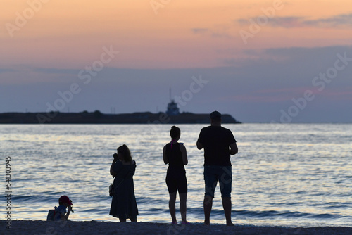 A family of four watches the sunset with a cape and a lighthouse in the background. Family members in the foreground. Silhouettes. Being on the beach of the island of Majorca. Selective focus. Relax.