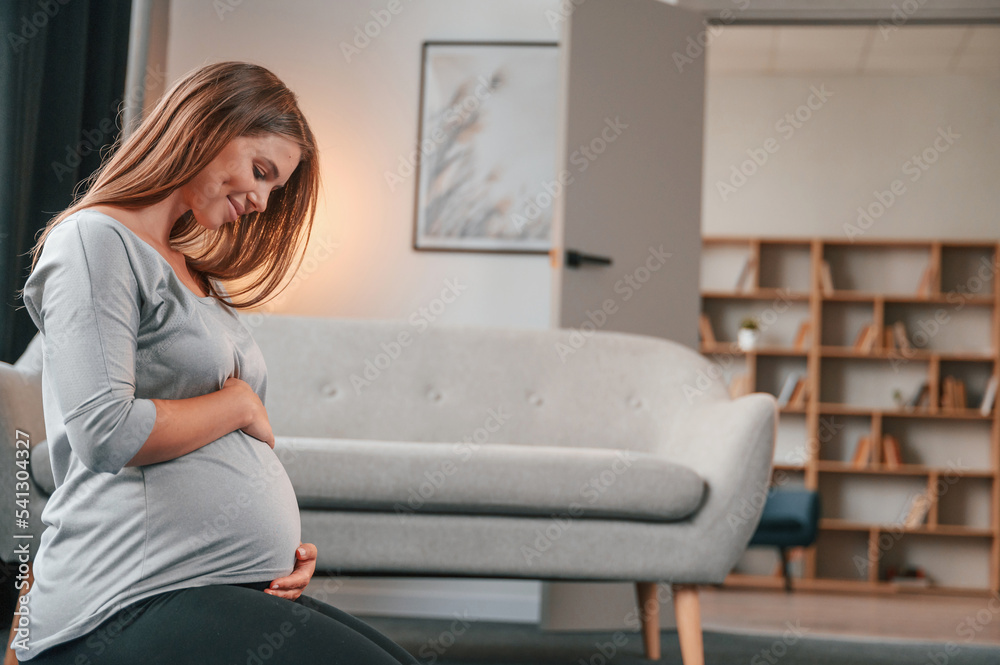 Side view. Sitting on the floor. Beautiful pregnant woman is indoors at home