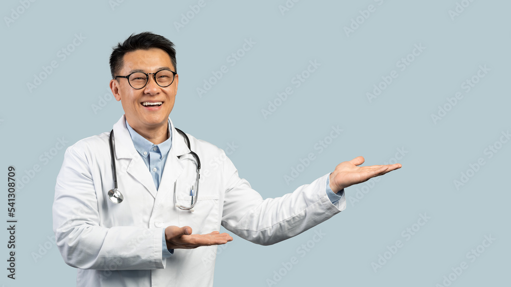 Cheerful mature chinese man doctor in white coat, glasses point hands to empty space
