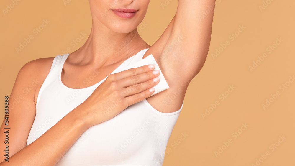 Hyperhidrosis concept. Closeup of young lady wiping her under arm area with napkin towel, standing on beige background Hyperhidrosis concept. Closeup of young lady wiping her under arm area with napkin towel, standing on beige background