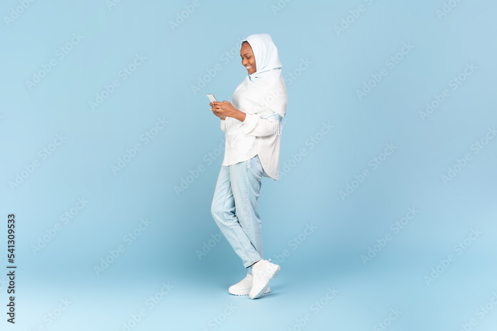 Happy black muslim woman texting on cellphone, communicating online while standing over blue background, full length Happy black muslim woman texting on cellphone, communicating online while standing over blue background, full length