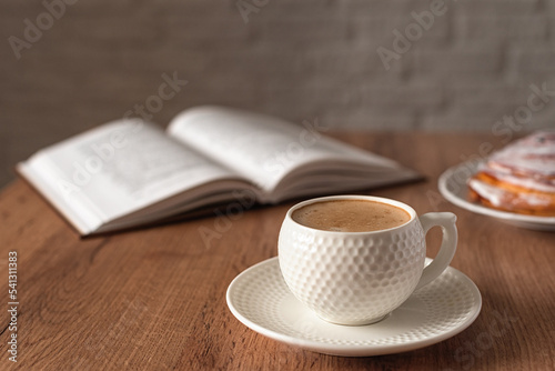 A concept for wishing a productive day. A cup of fragrant cappuccino, a cinnabon bun and a book on the wooden kitchen table. Still life in the style of minimalism. Selective focus. Space for text.