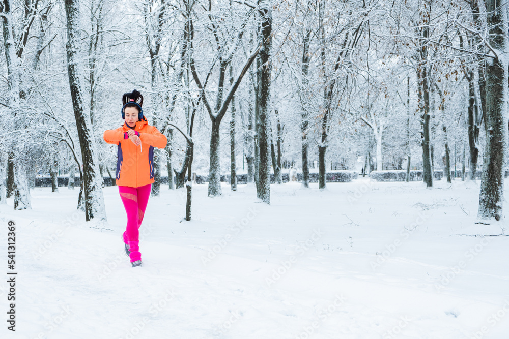 Running woman looking at smartwatch, checking heartrate and pulse during jogging in winter park. Fitness woman setting up smart watch for running training during winter workout