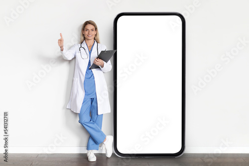 Medical App. Smiling Doctor Lady In Uniform Posing Near Big Blank Smartphone
