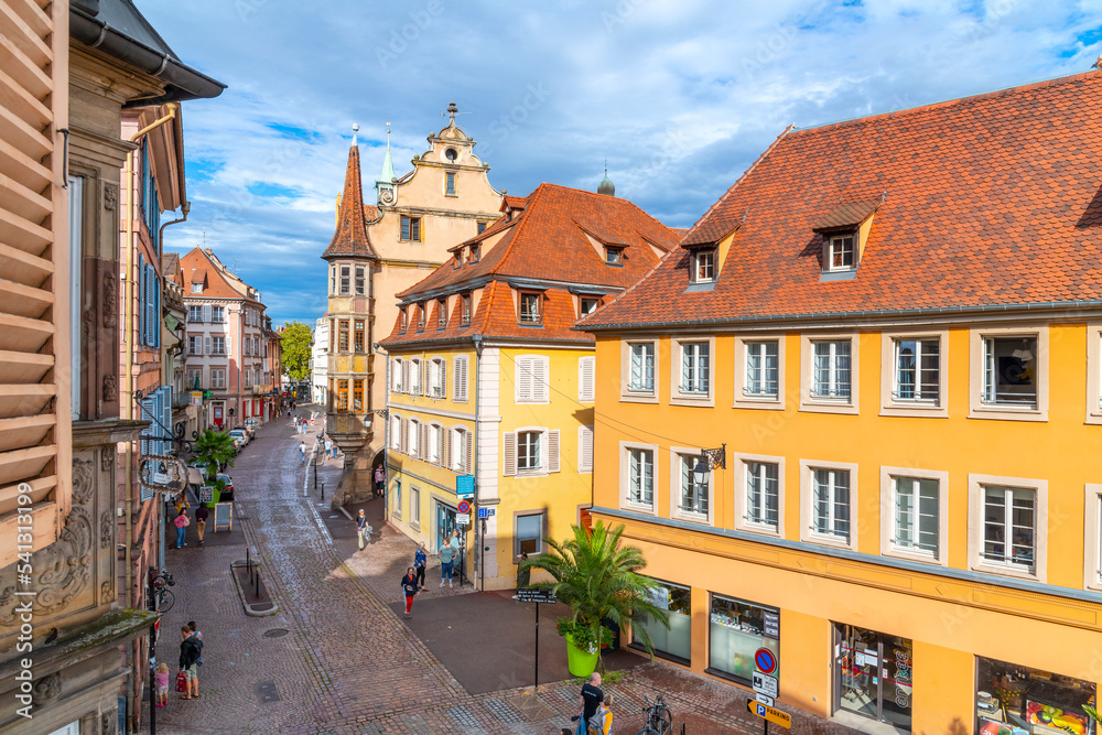 View from a window of a picturesque street of shops and sidewalk cafes in the medieval old town of Colmar, France, one of the stops along the wine route in the Alsace region. View from a window of a picturesque street of shops and sidewalk cafes in the medieval old town of Colmar, France, one of the stops along the wine route in the Alsace region.