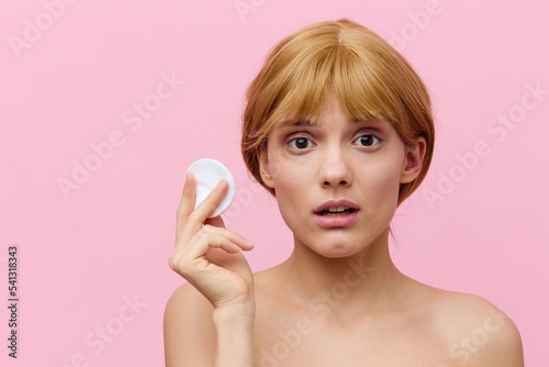 a shocked, surprised woman stands on a pink background holding a cotton pad in her hand, looking into the camera with her blonde hair pinned up from behind. Horizontal photo with empty space