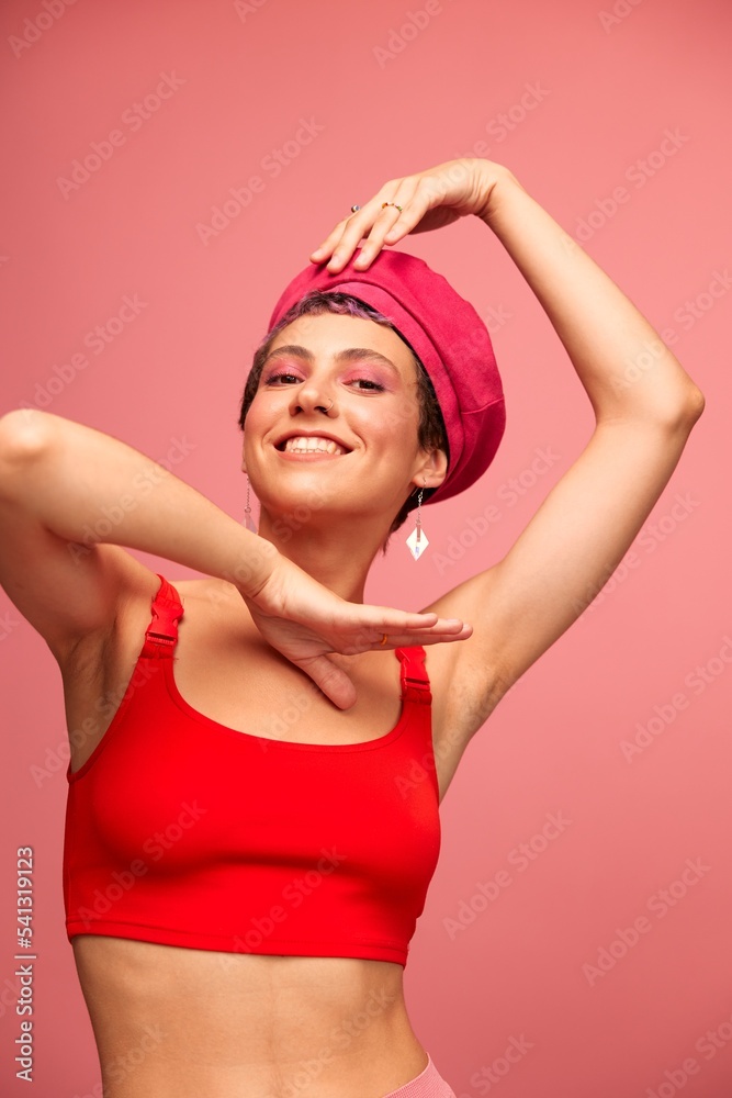 Young athletic woman with a short haircut and purple hair in a red top and a pink hat with an athletic figure smiles and grimaces looking at the camera on a pink background Young athletic woman with a short haircut and purple hair in a red top and a pink hat with an athletic figure smiles and grimaces looking at the camera on a pink background