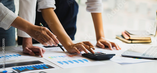 Working as a team with analytical graphs, business people are using calculators to calculate investment, turnover and profits displayed in a document on a desk at a conference room.