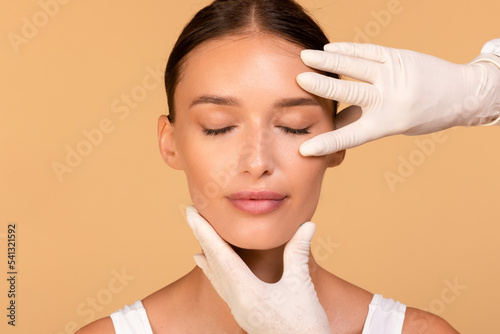 Facial treatment. Cosmetologist touching beautiful caucasian woman face, lady sitting over beige background photo
