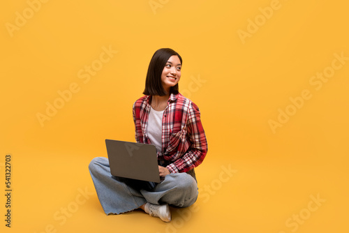Happy asian lady sitting on floor with new modern laptop, looking aside at free space and smiling over yellow background
