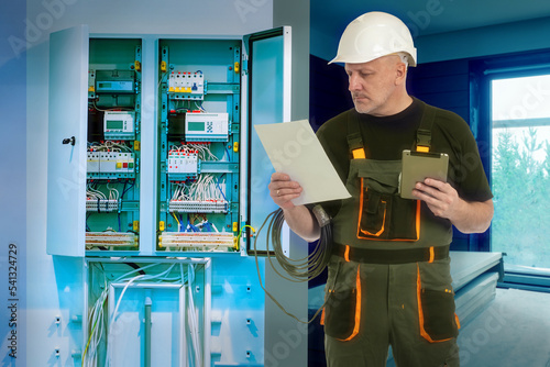 Electrical technician at work. Man with tablet and sheet paper. Electrical technician near switchboard. Setting up power grids in house under construction. Electrical technician man in white helmet