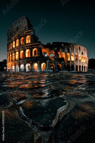 Portrait picture of the Roman Colosseum at night with wet cobblestones reflecting the illuminated ancient arena