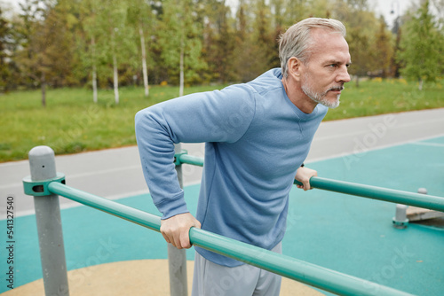 Side view portrait of active mature man exercising on parallel bars outdoors, copy space
