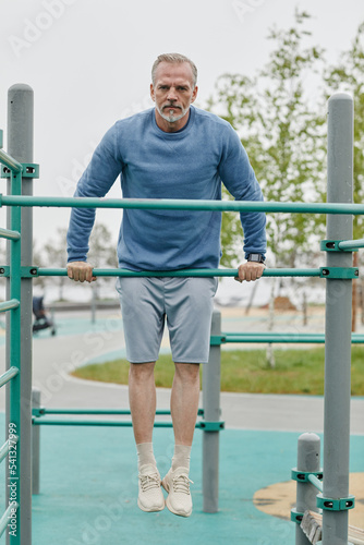 Full length portrait of sportive mature man exercising on parallel bars outdoors and looking at camera