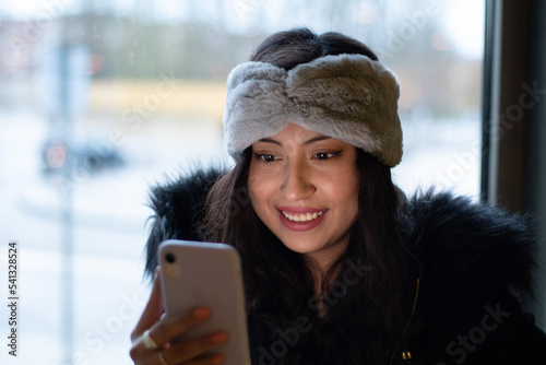 Latina girl is shopping in a mall, she looks at her cell phone and smiles she is next to a window facing outside.