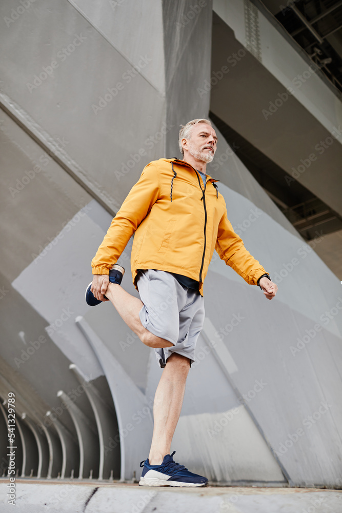 Vertical full length shot of senior man stretching legs outdoors while arming up for jogging