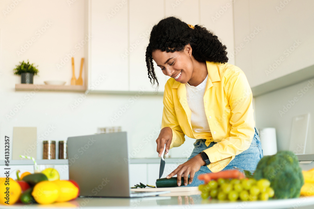 Cheerful black lady cutting organic vegetables and using laptop, cooking food in minimalist kitchen interior, free space