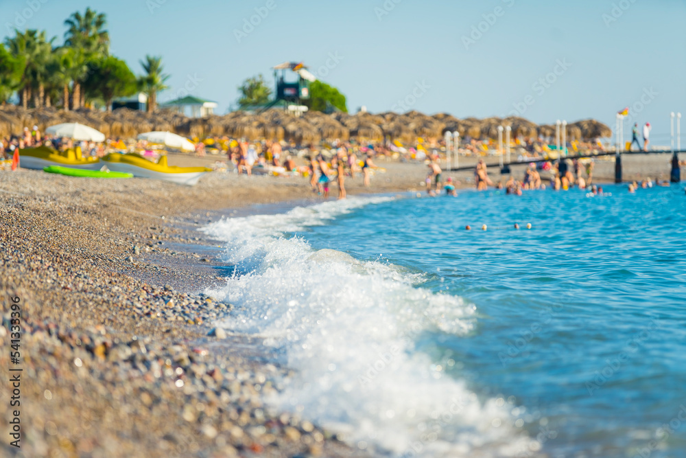 Summer holidays on a pebble beach. Turquoise seawater touching the pebble beach. Blurred tourists silhouettes in the background. Okurcalar, Turkey. High quality photo