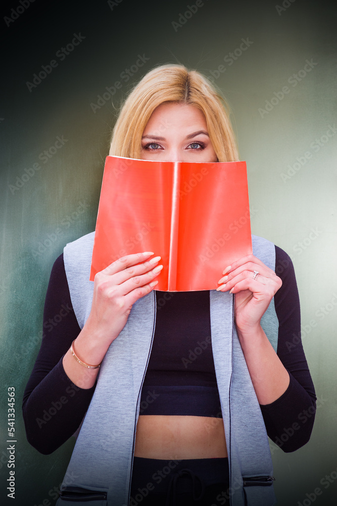 I love book. Blonde Eastern European Woman holding opened book, covering face, thinking, wearing long vest, black fitted T shirt, standing by metal silver wall in New York. .
