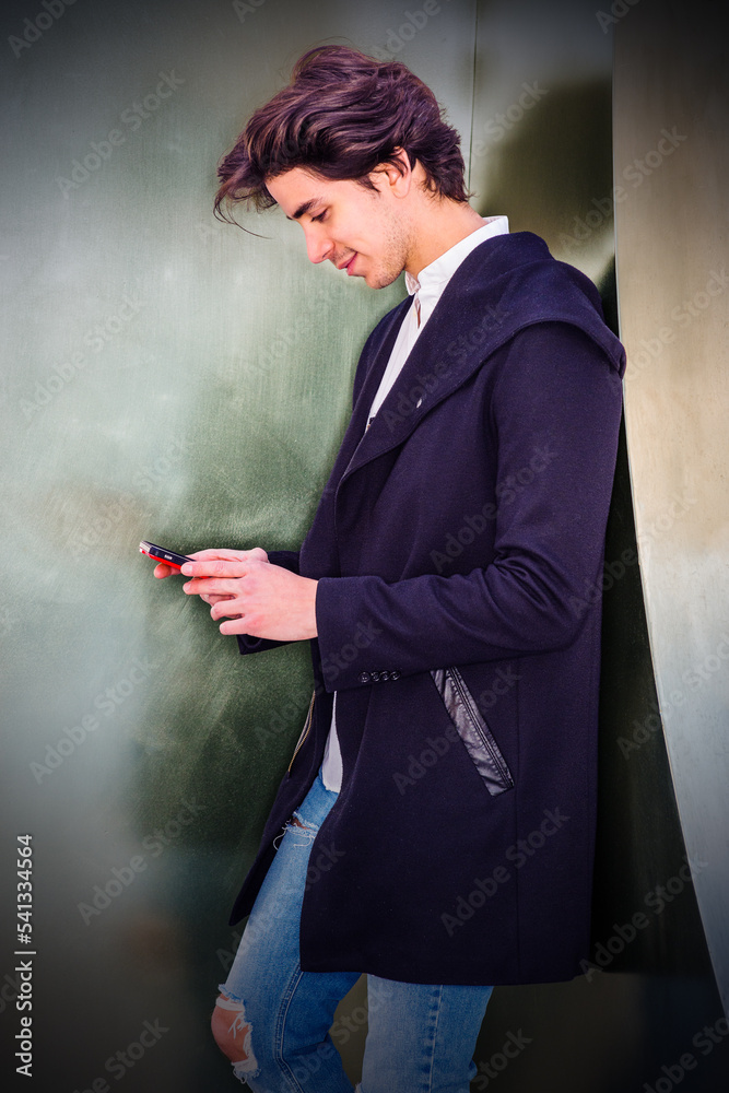 Texting anywhere. American college student wearing fashionable long coat, jeans, standing against metal wall in New York, holding cell phone, looking down, reading messages..
