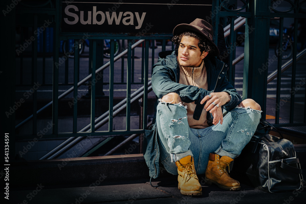 Traveling in New York City. Young Man wearing blue jacket with hood, jeans, yellow boot shoes, Fedora hat, black leather bag on ground, sitting on street by Subway sign.