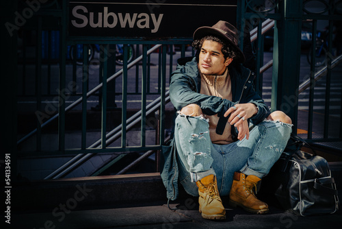 Traveling in New York City. Young Man wearing blue jacket with hood, jeans, yellow boot shoes, Fedora hat, black leather bag on ground, sitting on street by Subway sign.