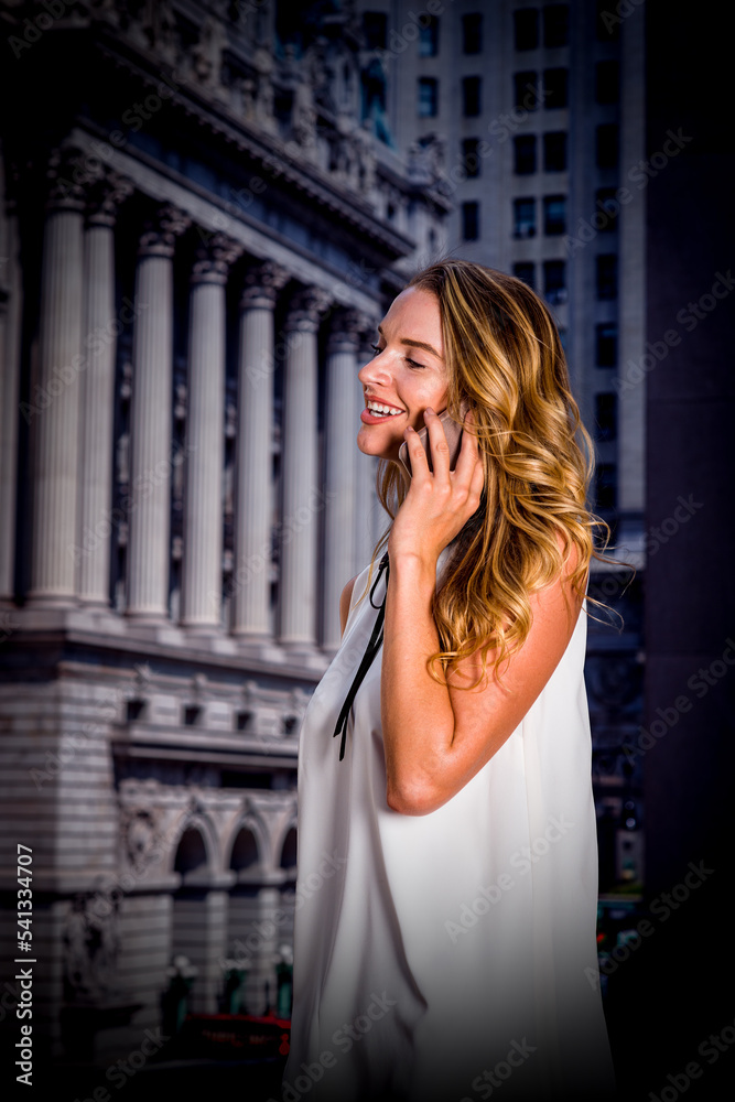 Young American Woman with blonde hair, wearing sleeveless white dress, standing in front of office building on street in New York City, talking on cell phone, smiling..