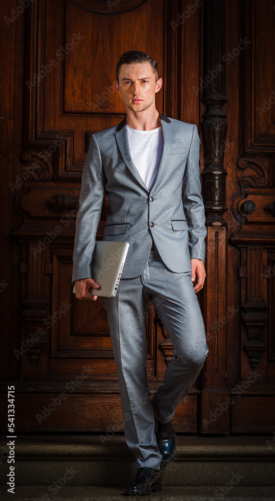 Young Eastern European Businessman working in New York City, wearing gray fashionable suit, black leather shoes, carrying laptop computer, walking down stairs of office doorway. .