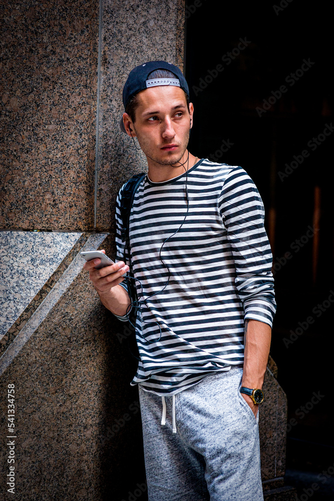 Young man wearing striped long sleeve T shirt, shoulder carrying bag, wearing cap backward, standing by column on street in New York City, texting on cell phone, listening music with earphone..