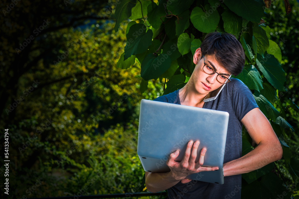 Young Asian American college student studying in New York City, standing by green big leaf plants on campus, works on laptop computer, talks on cell phone held between head and shoulder. .