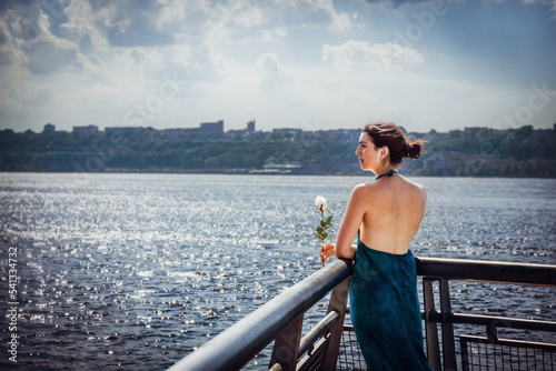 Obraz na płótnie Waiting in the Summer. Young woman with bare back, long blue dress, holding white rose, standing by fence by Hudson River in New York City, looking far away.. w salonie