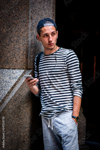Young man wearing striped long sleeve T shirt, shoulder carrying bag, wearing cap backward, standing by column on street in New York City, texting on cell phone, listening music with earphone..