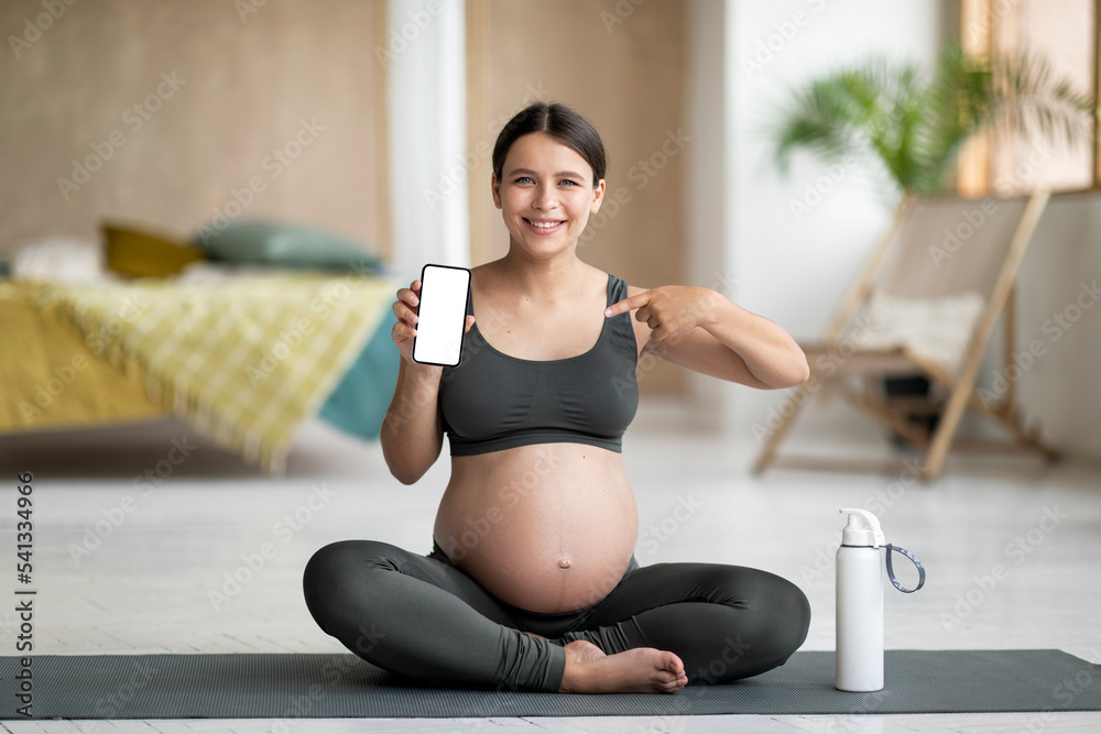 Fitness App. Pregnant Woman Sitting On Yoga Mat And Showing Blank Smartphone
