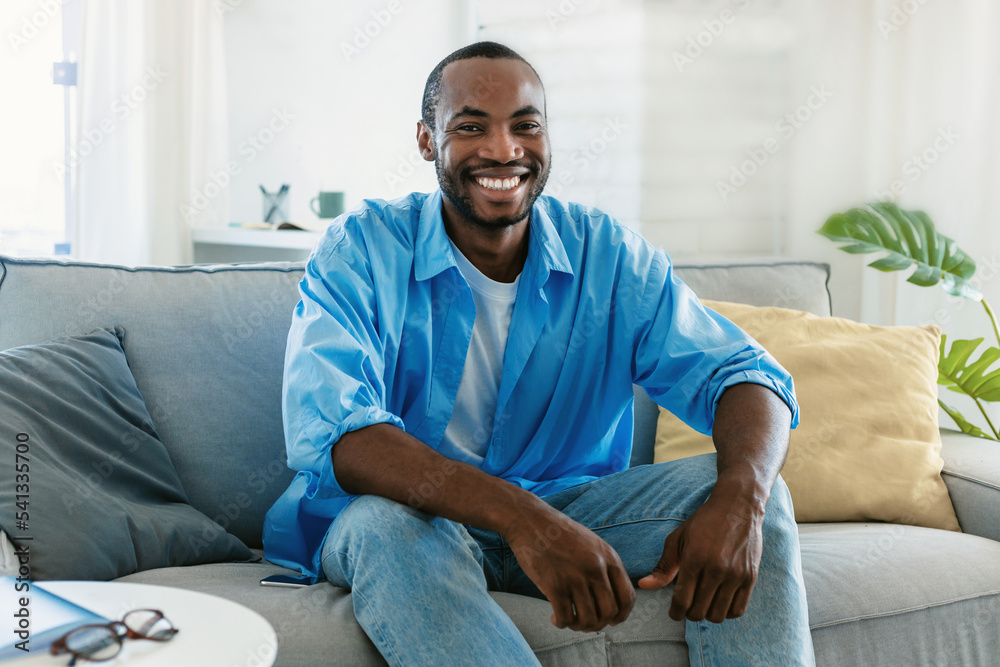 Portrait of handsome black man smiling and looking at camera, sitting on sofa at home, spending time indoors
