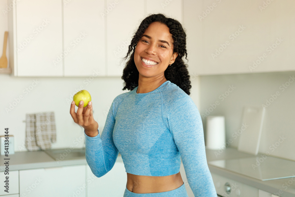 African american sporty lady holding apple and smiling at camera, eating fresh fruit after workout, standing in kitchen African american sporty lady holding apple and smiling at camera, eating fresh fruit after workout, standing in kitchen