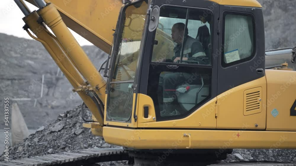 An excavator works at a gold mining site. The excavator cabin and the operator in it are shown.
