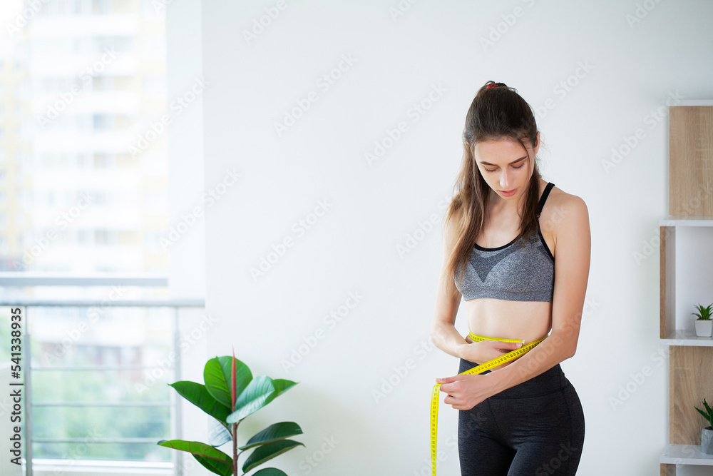 Slim young woman measuring her thin waist with a tape measure Slim young woman measuring her thin waist with a tape measure