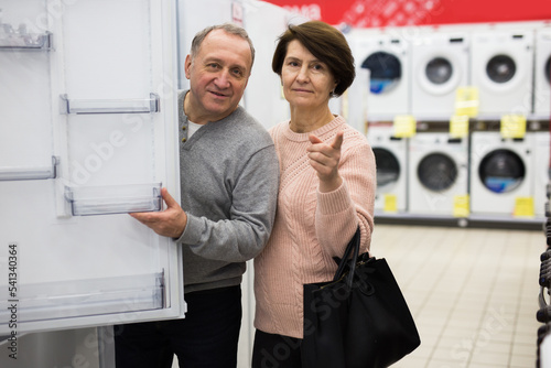 Senior couple choosing refrigerator while shopping in appliance store. Woman pointing finger.