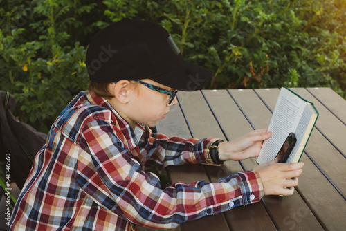 A teenager after school sits in a park and hides his phone in a book, pretends to read, while he looks at his phone