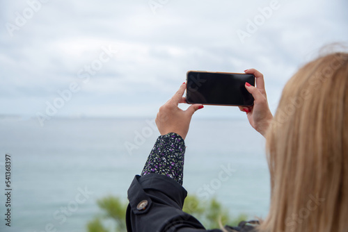 Mujer joven rubia con uñas rojas sacando una foto al paisaje  con dos manos con su teléfono móvil en horizontal durante un día nublado en la playa de El Sardinero 
