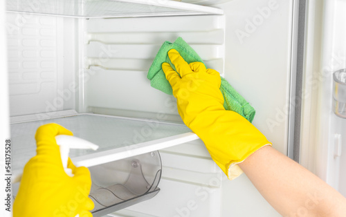 Deep Cleaning service. Woman hand in yellow rubber protective glove cleaning white open empty refrigerator with green rag. Spray for windows and glass surfaces cleaner