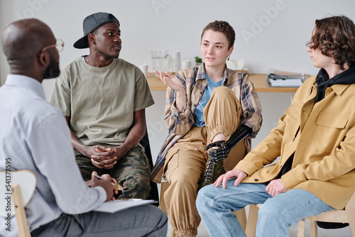 Girl sitting on chair and talking to psychologist at psychology class with other young people
