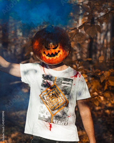 Man wearing pumpkin on his head in a park during fall with blue smoke in the background