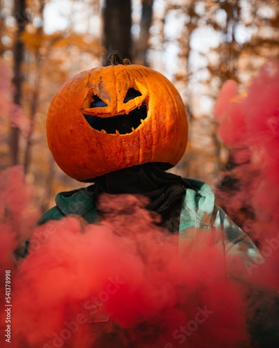 Person wearing pumpkin on his head in a park during fall with red smoke around him