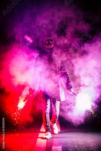 Male person wearing the scary mask and holding red-purple light fireworks on his hands, vertical