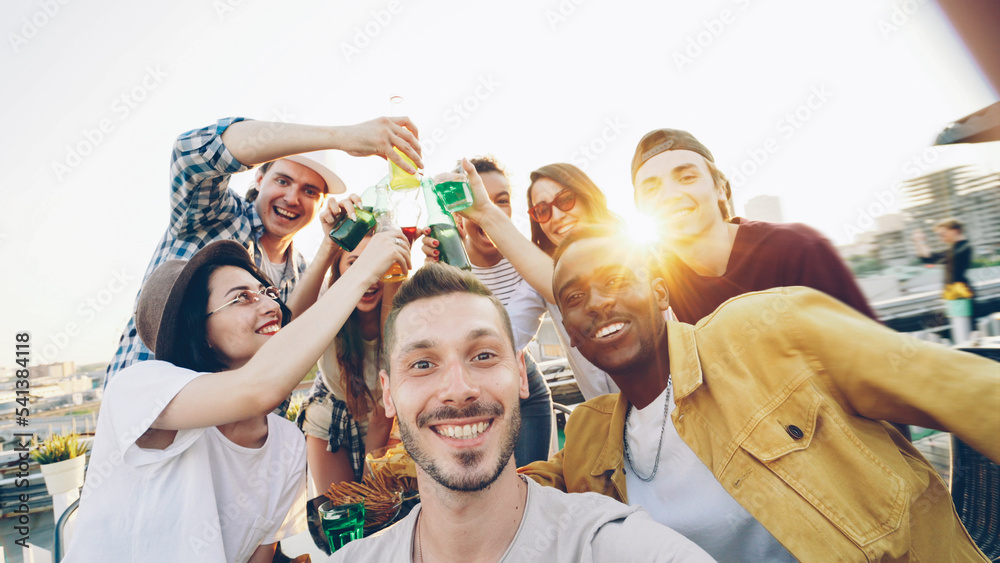 Obraz premium Point of view shot of handsome guy holding camera and taking selfie while his friends having fun