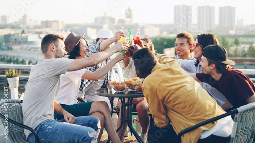 Multiethnic group of friends is talking, toasting then clanging bottles and glasses and drinking beer and soft drinks at table on roof. Beautiful view of big city is in background.