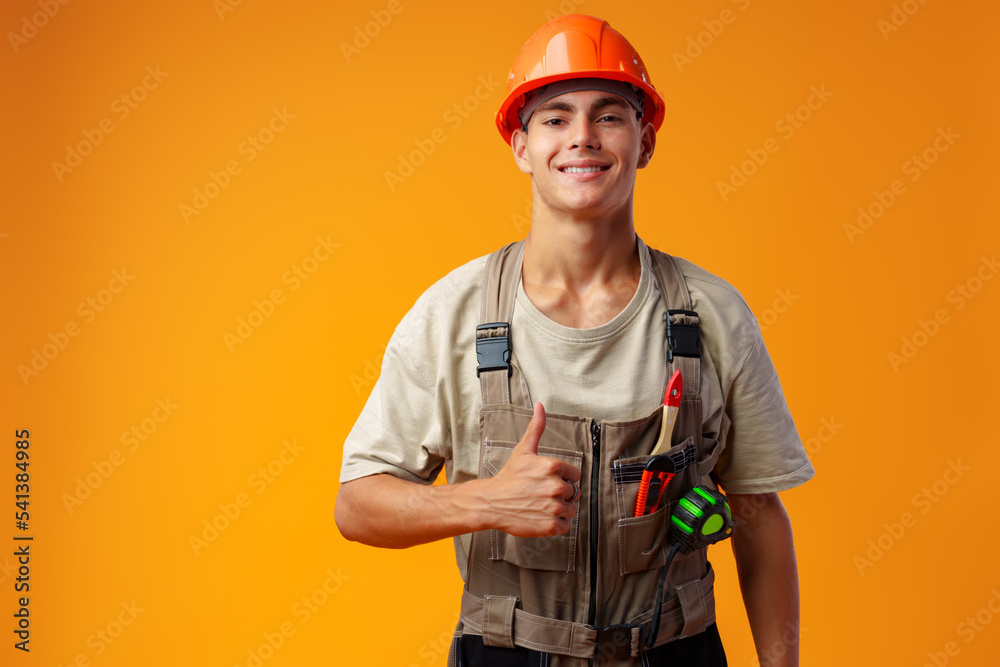 Young construction worker in helmet and uniform posing on yellow background in studio Young construction worker in helmet and uniform posing on yellow background in studio